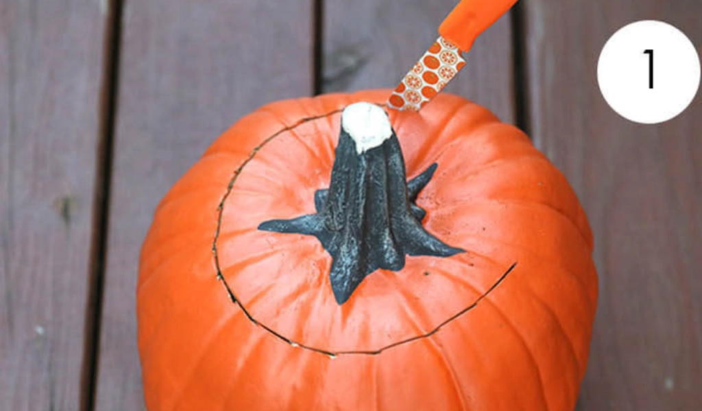 A knife is cutting a circular lid around the stem of an orange pumpkin placed on a wooden surface.