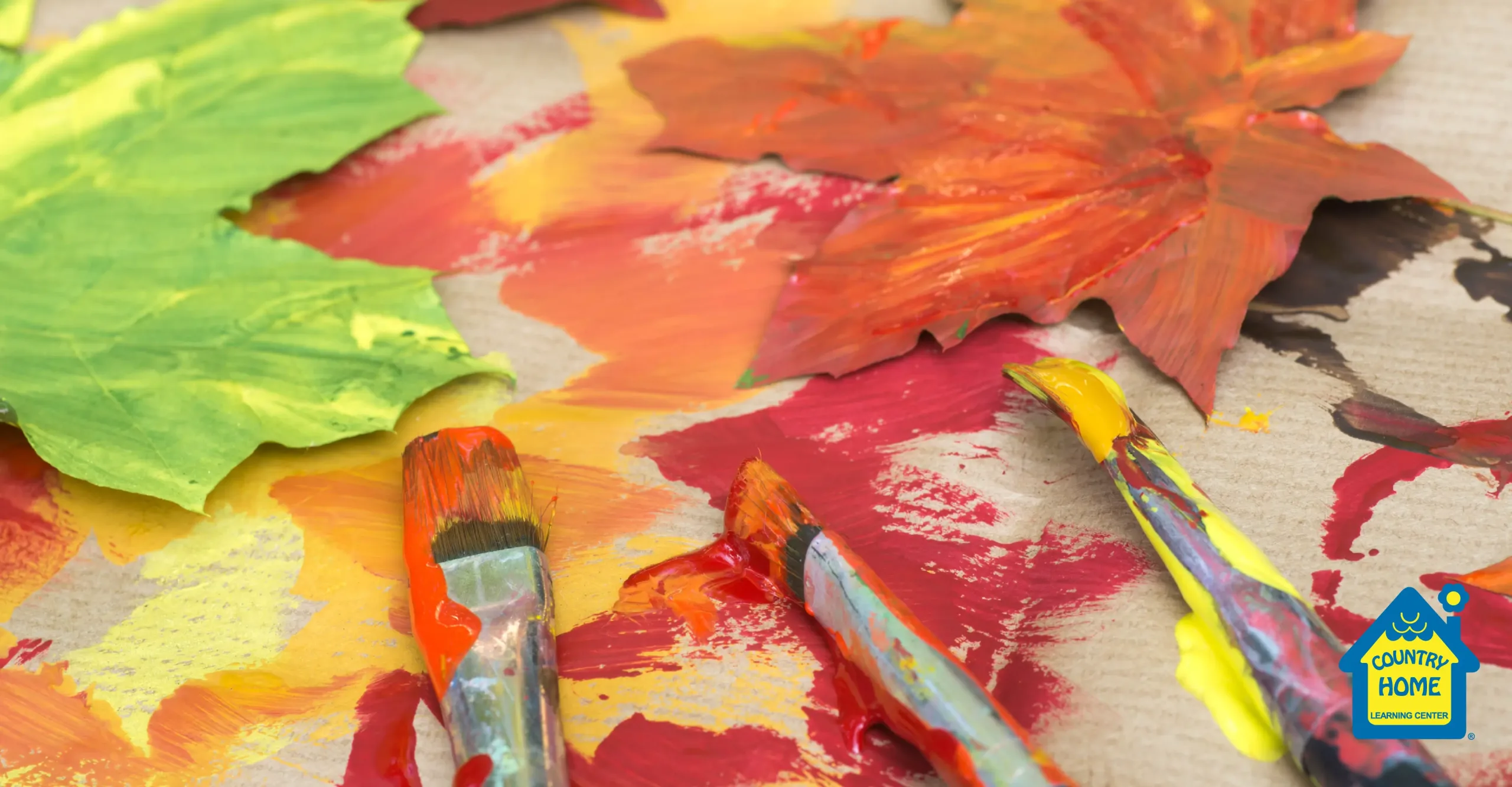 Three paintbrushes with red, yellow, and orange paint lie on a surface with painted paper leaves in autumn colors—perfect inspiration for fall crafts. The Country Home Learning Center logo is in the bottom right corner.