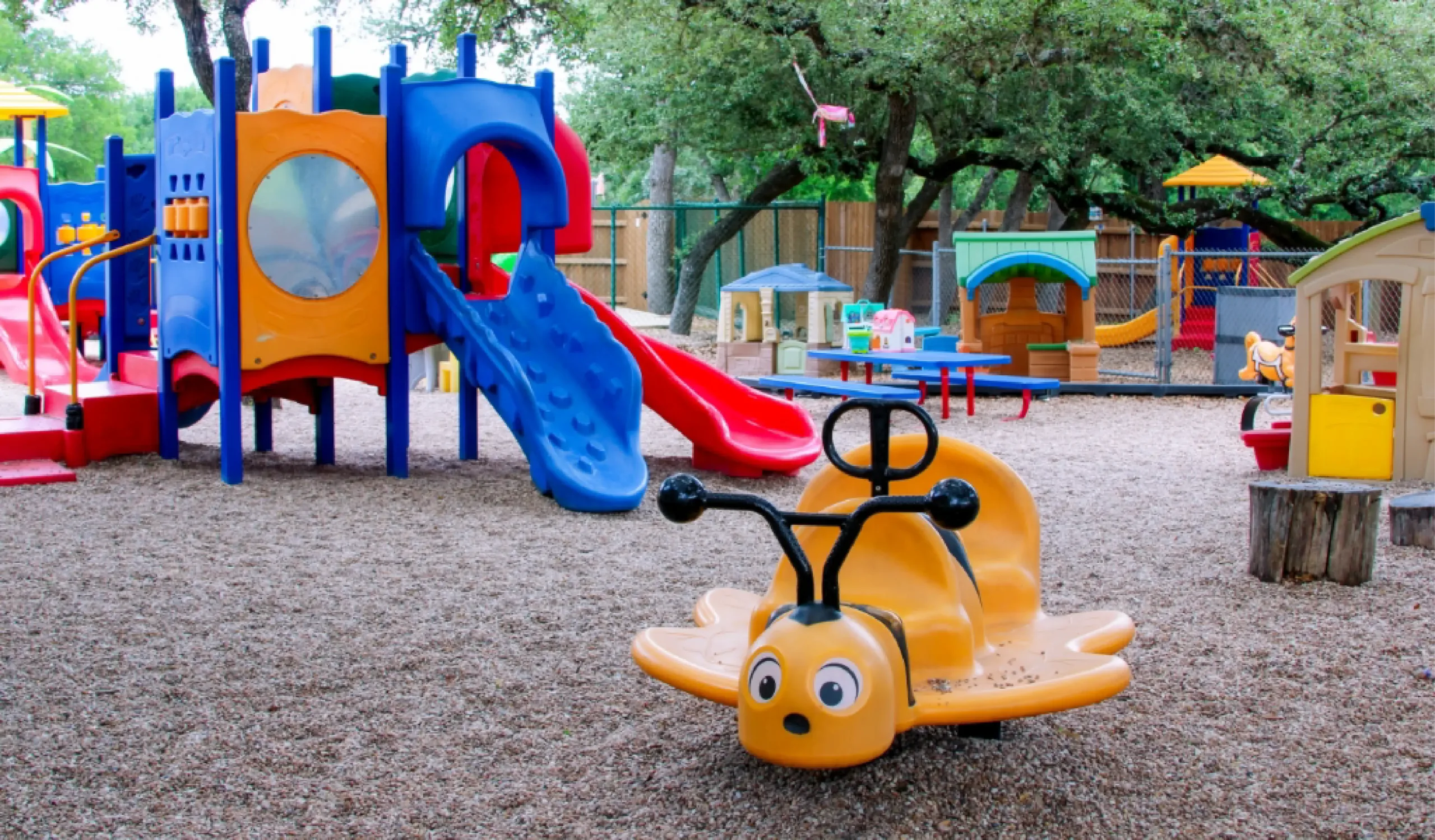 Outdoor playground with colorful climbing structures, slides, a yellow bee-shaped rocker, picnic tables, and trees in the background.