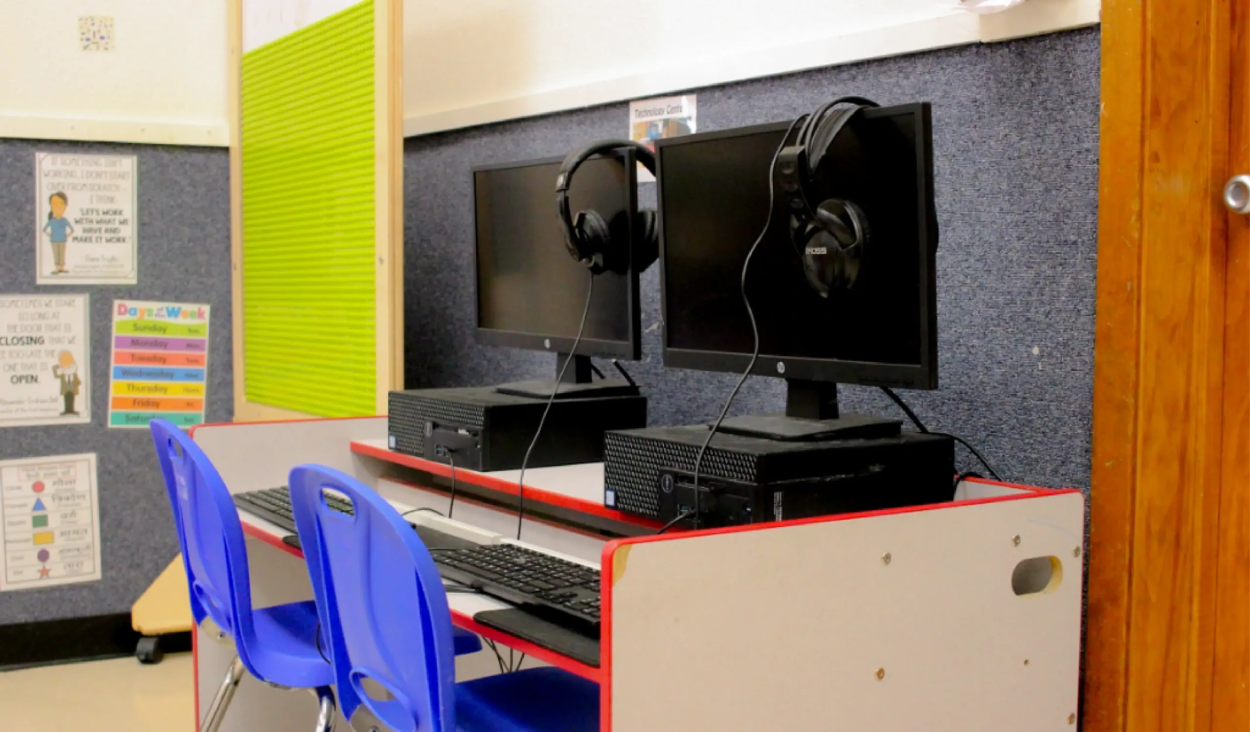 Two computer workstations with monitors, keyboards, and headphones are set up on a desk with two blue chairs in a classroom setting.