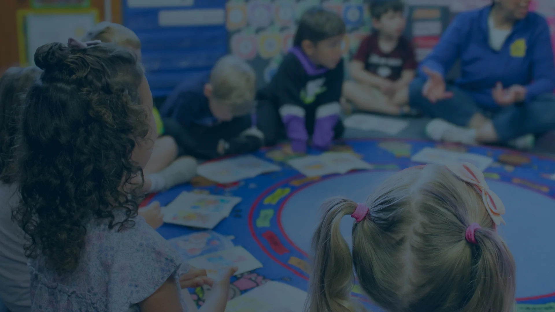 Children sit in a circle on a colorful classroom rug, looking at books, while an adult leads an activity.
