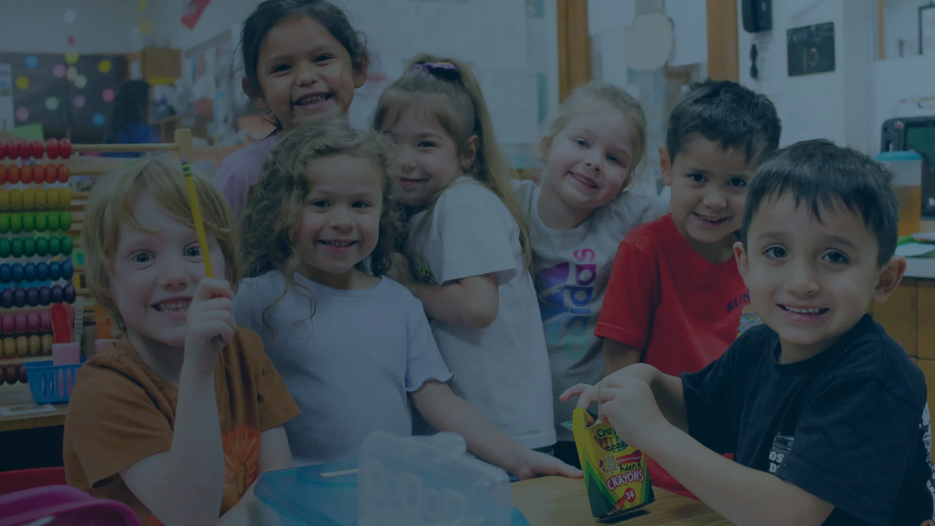 A group of seven young children sit and stand around a classroom table, smiling at the camera with school supplies visible.