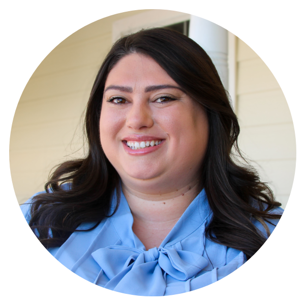 Teacher – 1604 & Shaenfield – 9 (Ms.Skye) A woman with long dark hair smiles at the camera while wearing a light blue blouse with a bow, standing in front of a light-colored wall at the daycare and preschool at Shaenfield Rd.