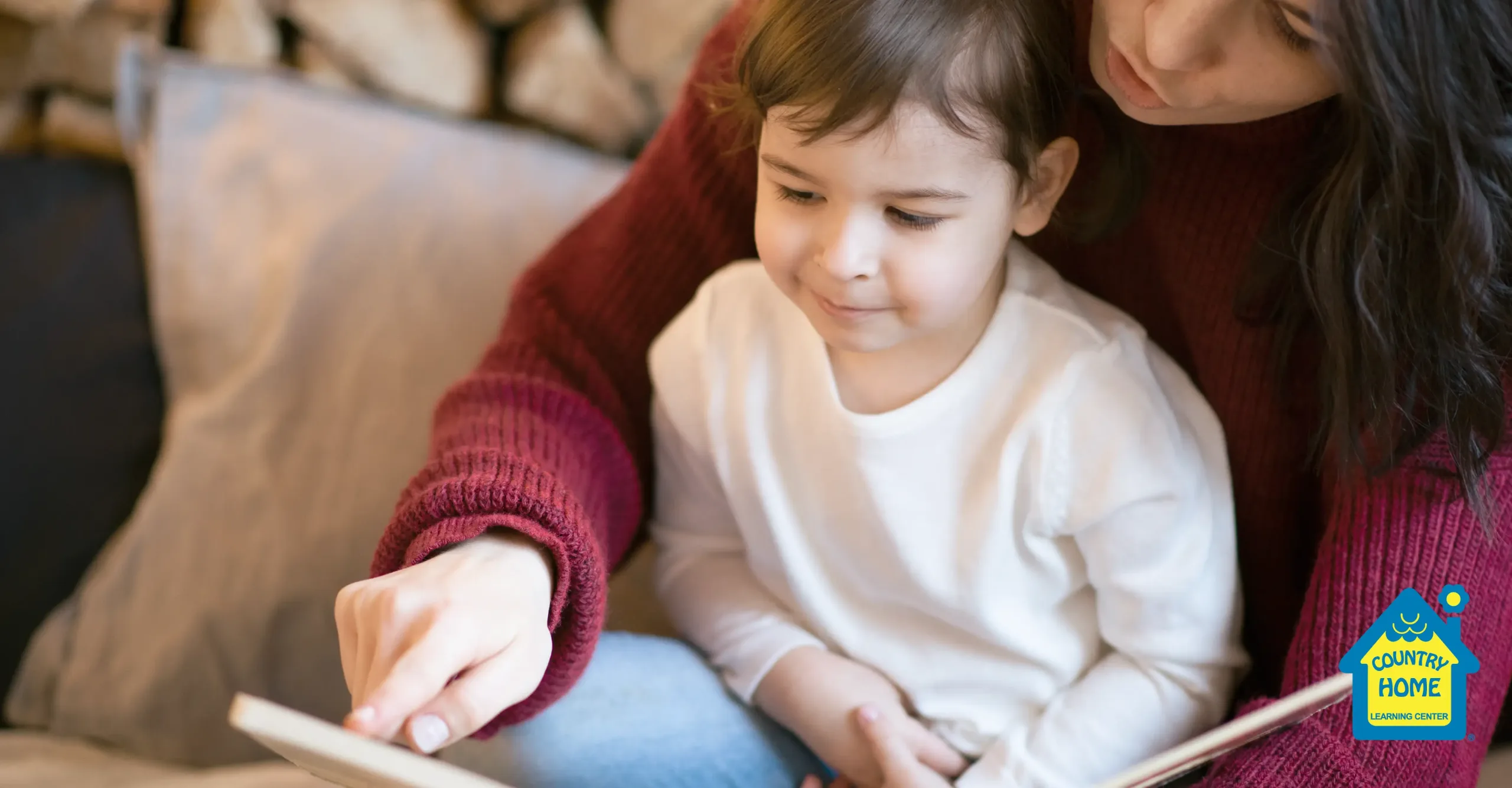 An adult and a young child sit together on a couch, reading a book. The adult points at the pages while the child looks on, fostering early childhood learning. A "Country Home Learning Center" logo appears in the corner.