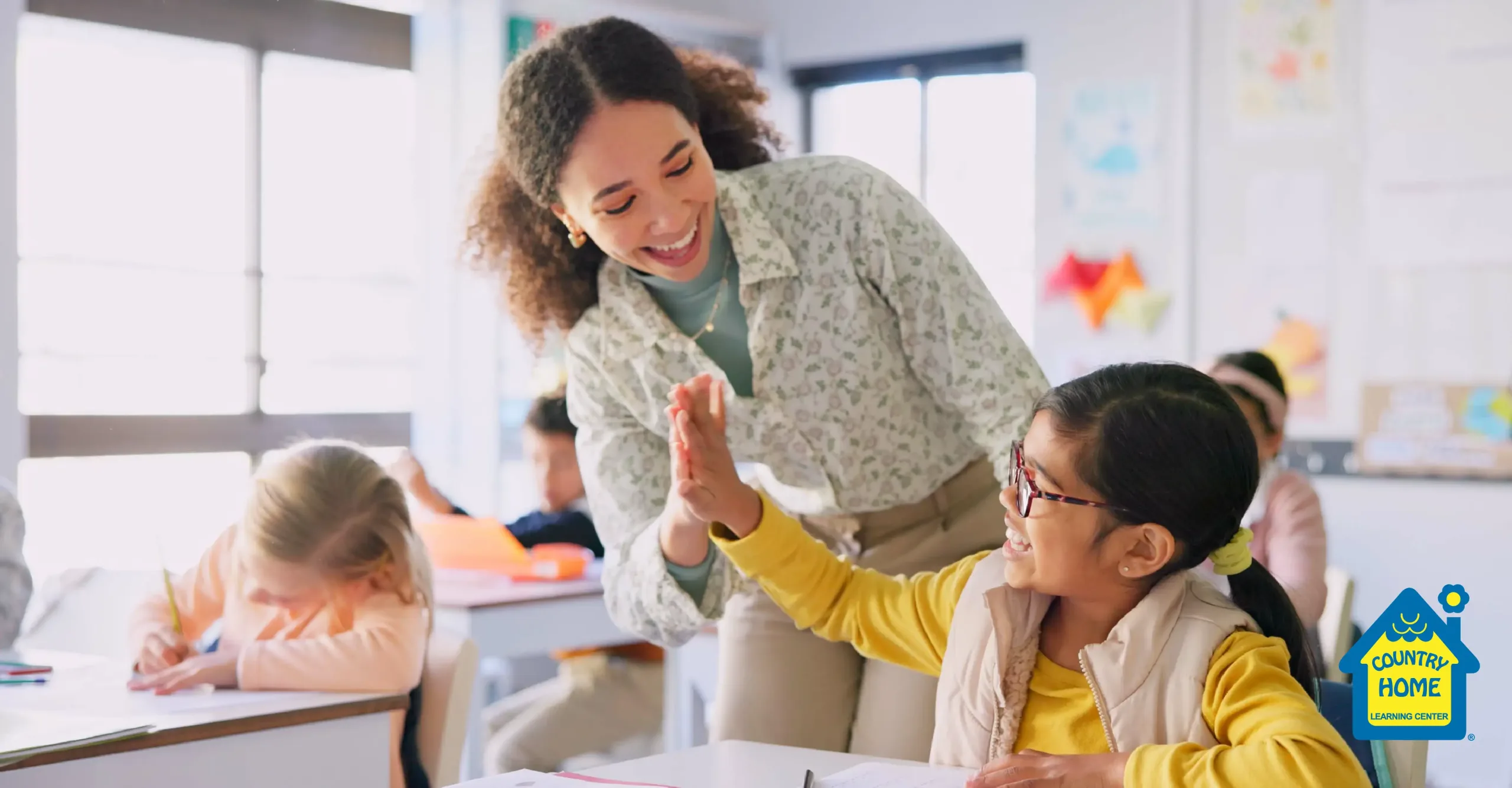 A teacher smiles and gives a high-five to an early childhood student wearing glasses in a classroom, while other students sit at their desks.