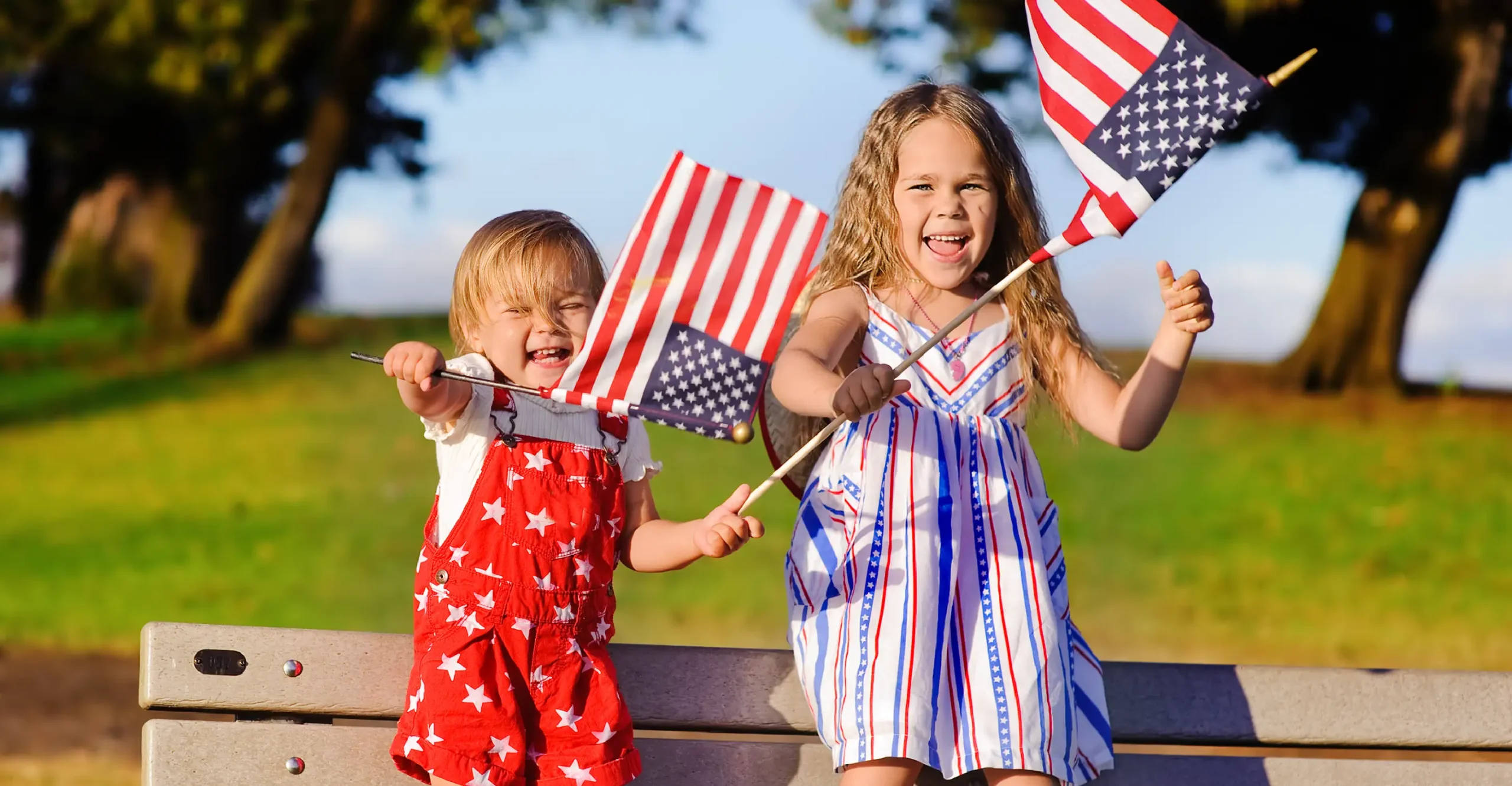 Two kids waving an American flag standing on a park bench.